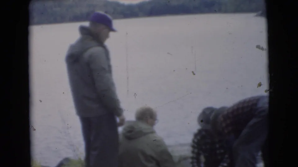 1956 Miami frame with a capped man standing at the lakeshore while others sit or bend near calm water.