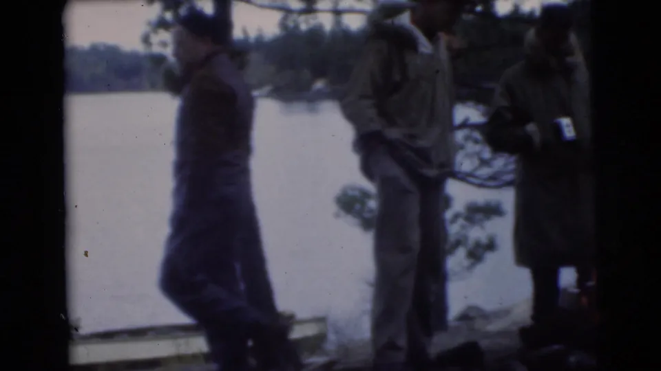 1956 Miami lakeside home-movie frame showing adult men in cold-weather clothing standing near water and trees.