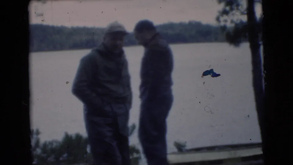 1956 Miami home-movie frame showing two adult men in coats standing close together at a cloudy lakeshore.
