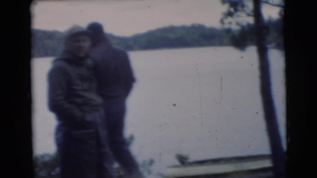 Two or more men stand near a lake, partly silhouetted and softly focused.