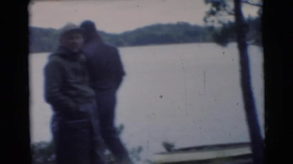 1956 Miami home-movie frame showing men in coats near a lakeshore, with trees and water behind them.