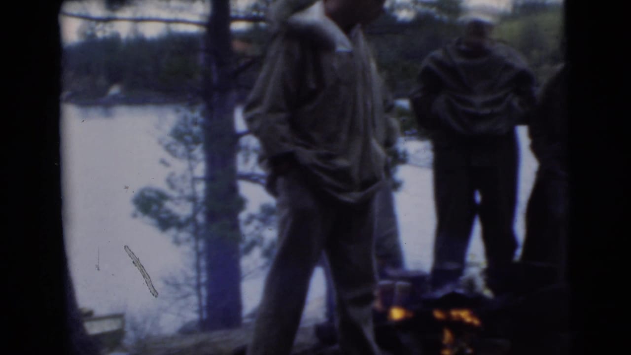 Men in coats stand near a small campfire with a lake and trees behind them.