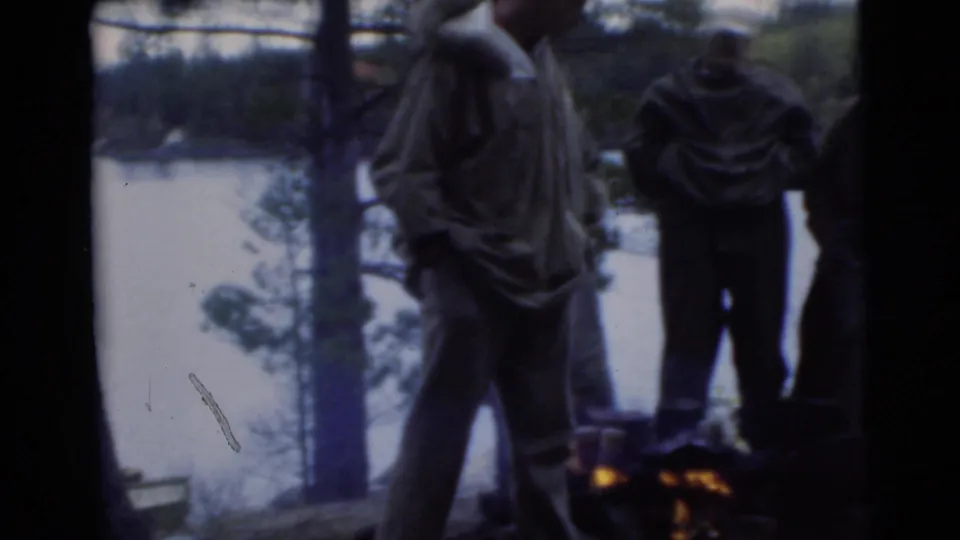 Adult men stand beside a small campfire near water in 1956 Miami archival 8mm footage.
