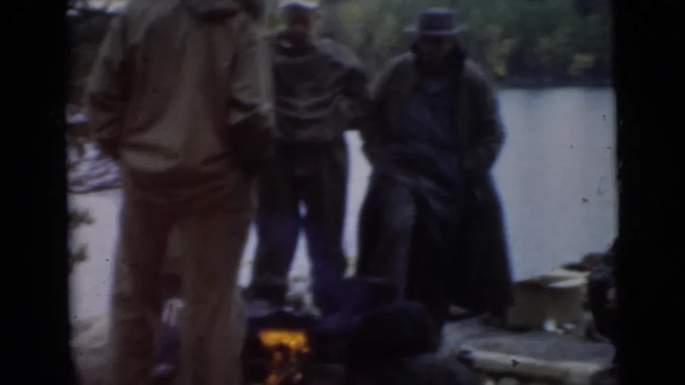 1956 Miami 8mm frame of men gathered around a small lakeside fire, with wooded shoreline behind them.