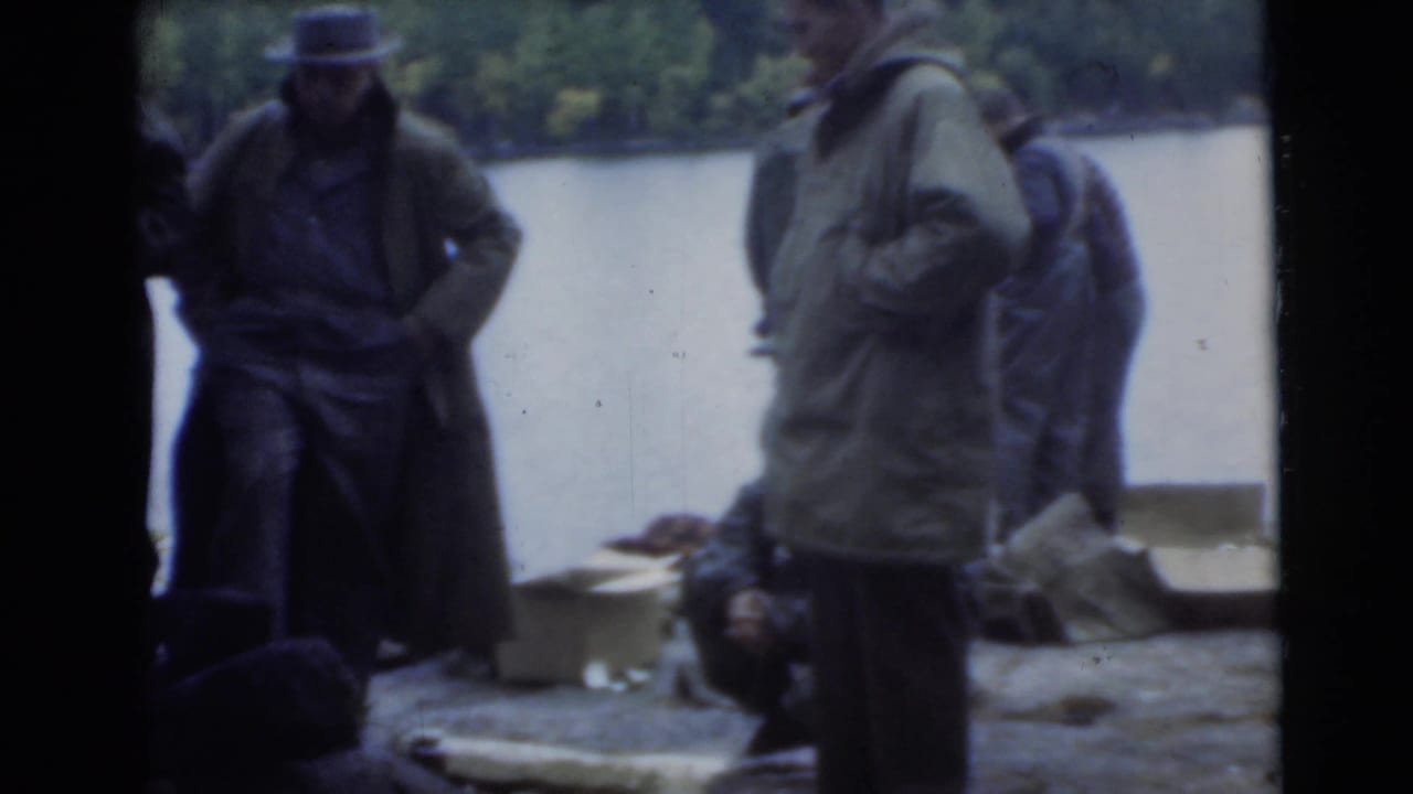 Men in coats and hats stand near water with gear on the ground.