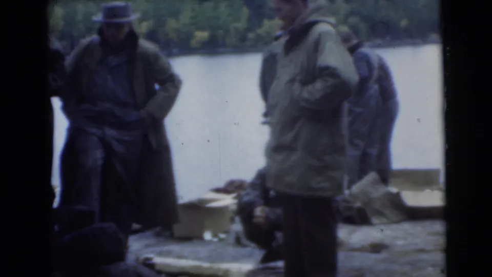 1956 Miami home-movie frame of bundled men standing near a lakeside area with bags or camping gear nearby.