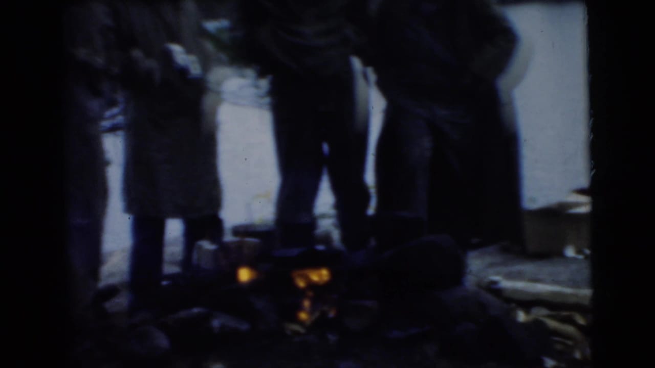 Dark silhouettes of men stand around a small glowing campfire.