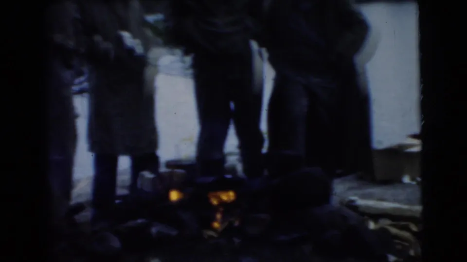 Dim 8mm frame from the Miami clip showing men in coats gathered around a small orange campfire near water.