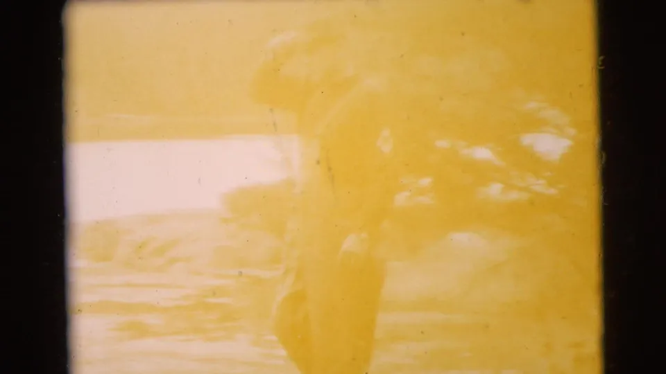 1956 Miami home-movie frame with a uniformed man faintly visible through yellow fading near a wooded waterfront.