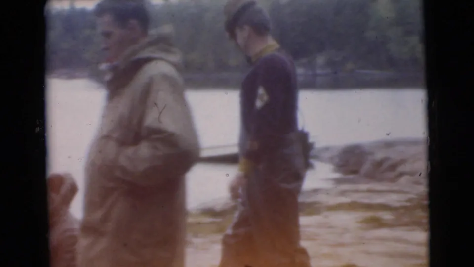 1956 Miami 8mm frame showing two adults walking by a shoreline, including a hooded figure and a man in uniform-like clothing.