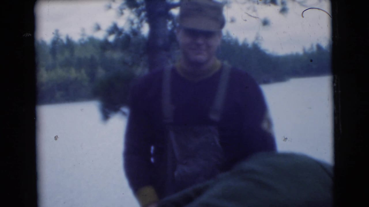 Man in cap and overalls stands near water with trees behind him.