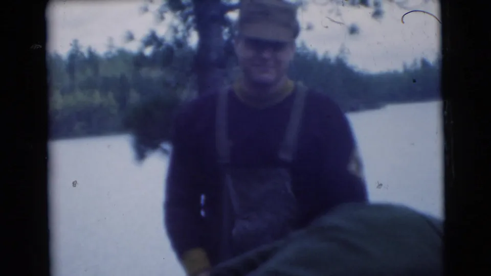In the 1956 Miami home-movie footage, a man wearing a cap and overalls stands by a lake with trees in the background.