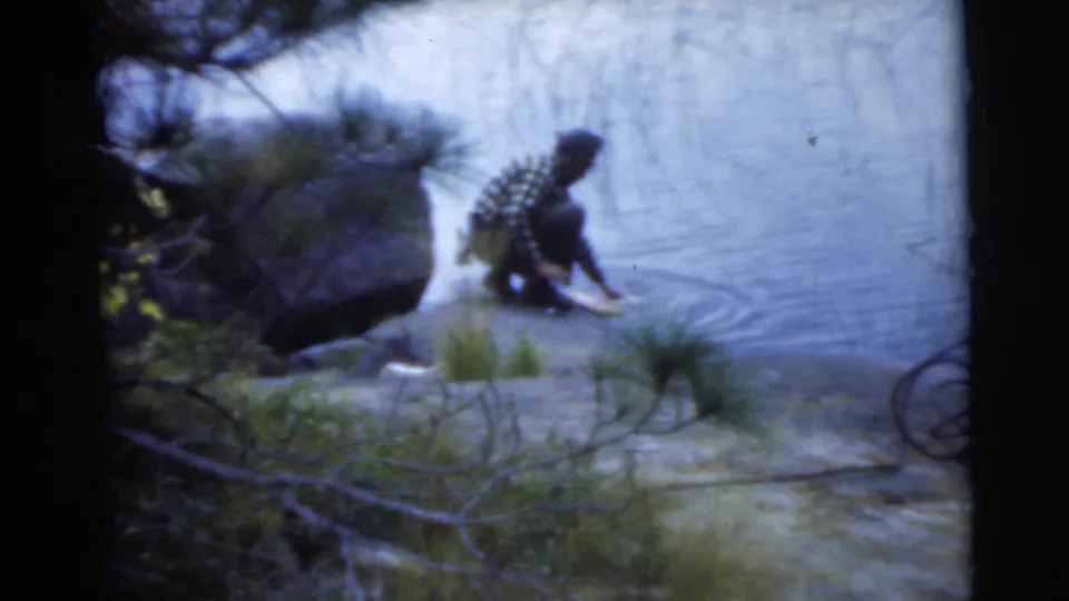 A man in a plaid shirt crouches at the edge of the lake, with shoreline rocks, water ripples, and trees visible in the 1956 Miami footage.