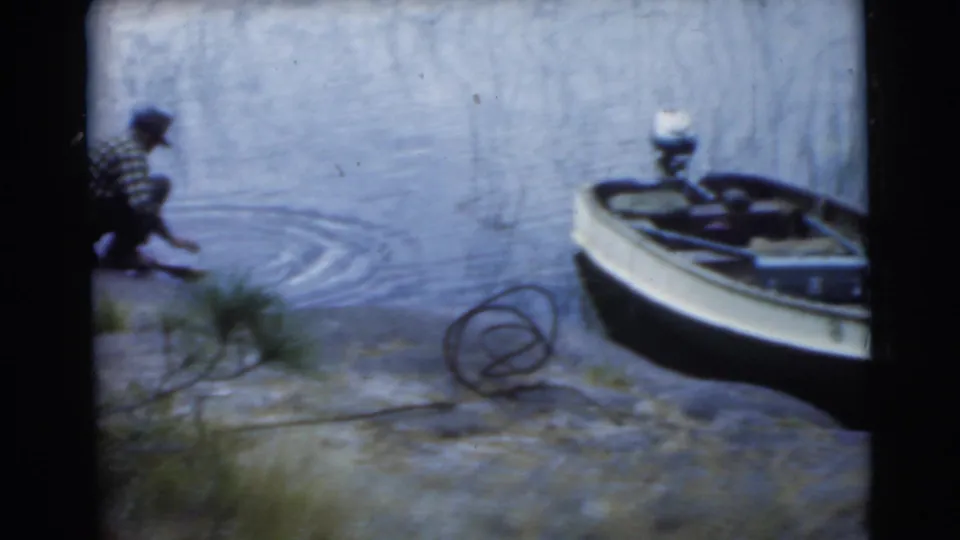A crouching man works at the lake edge while a small boat rests nearby along the Miami shoreline in the 1956 8mm home movie.
