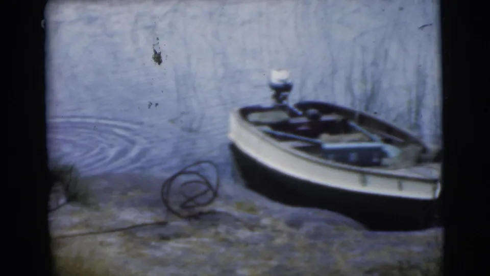 1956 Miami home-movie frame showing a small motorboat near a rocky lake edge with ripples and pine branches.
