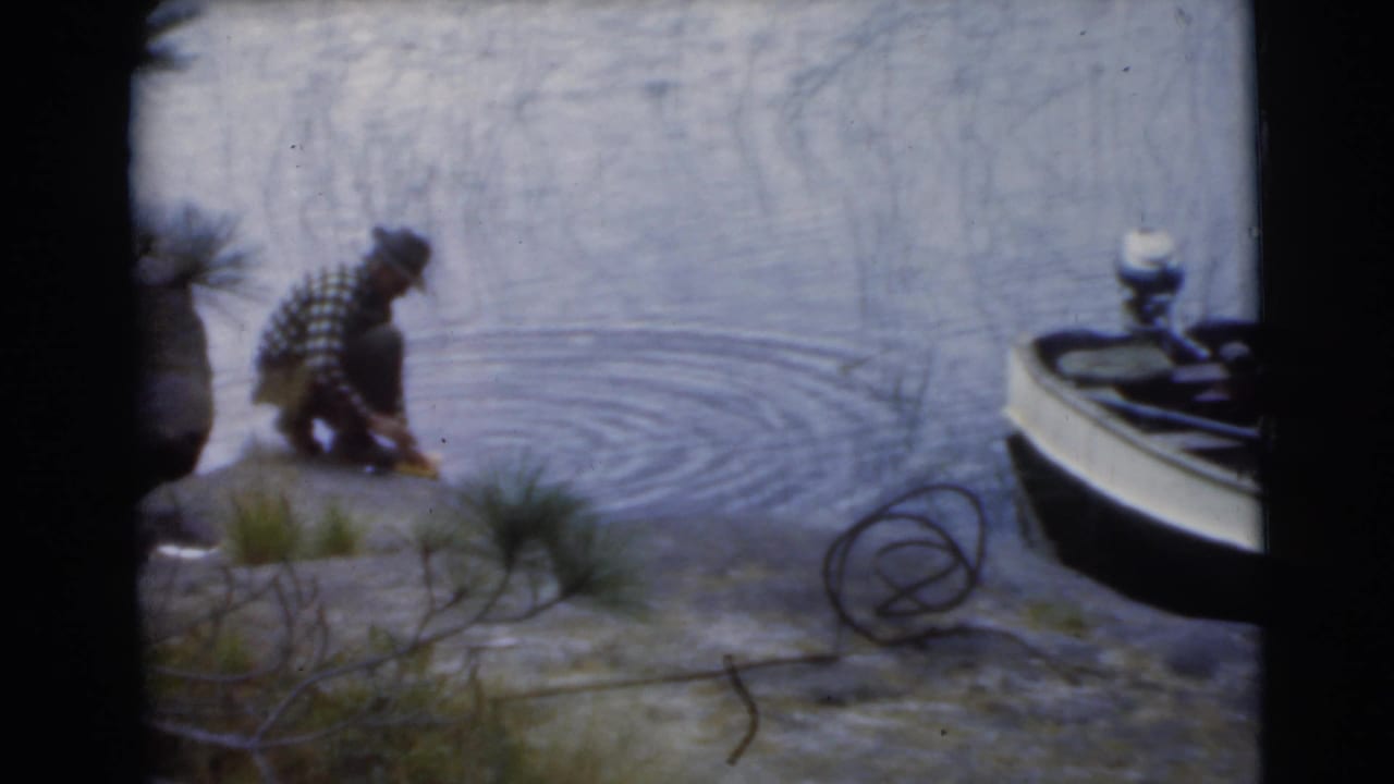 Man crouches by a lake edge near a small motorboat and rippling water.