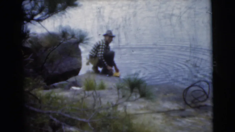 1956 Miami home-movie frame showing a crouching man at a rocky lake shore with rippling water nearby.