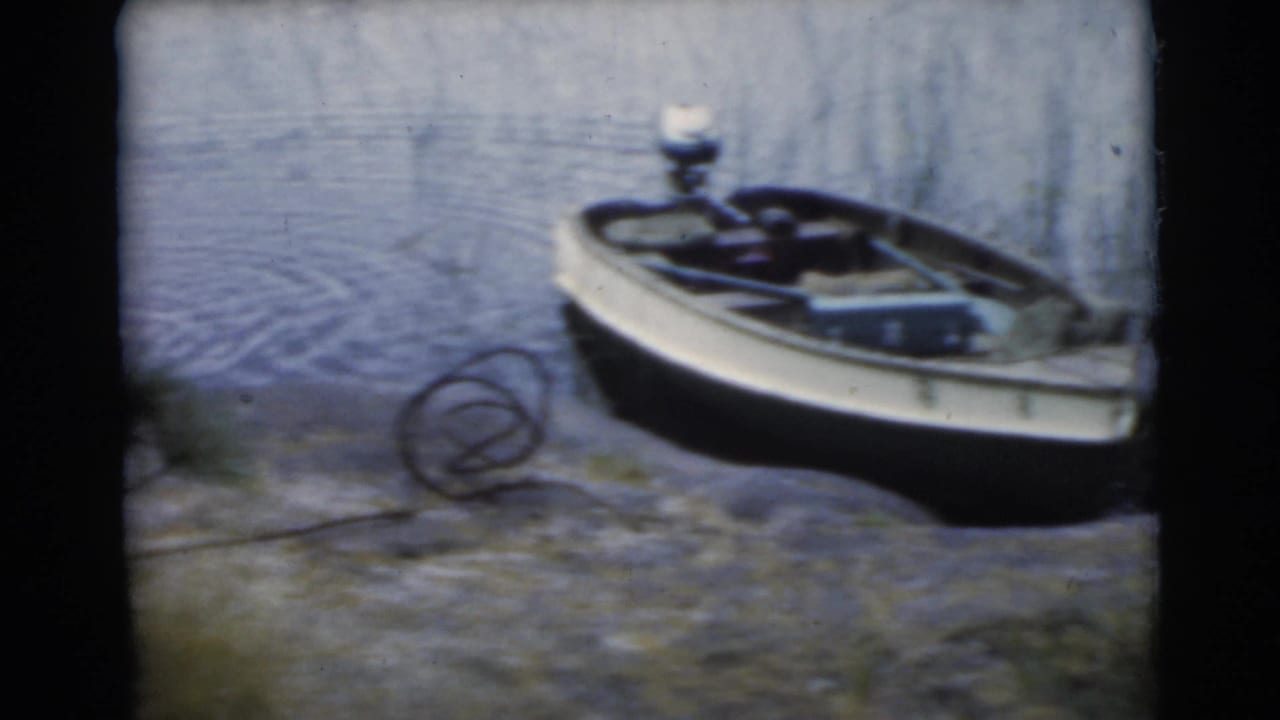 1956 8mm frame from Miami metadata showing a small boat at the lakeside with rope on the shore and water behind it.