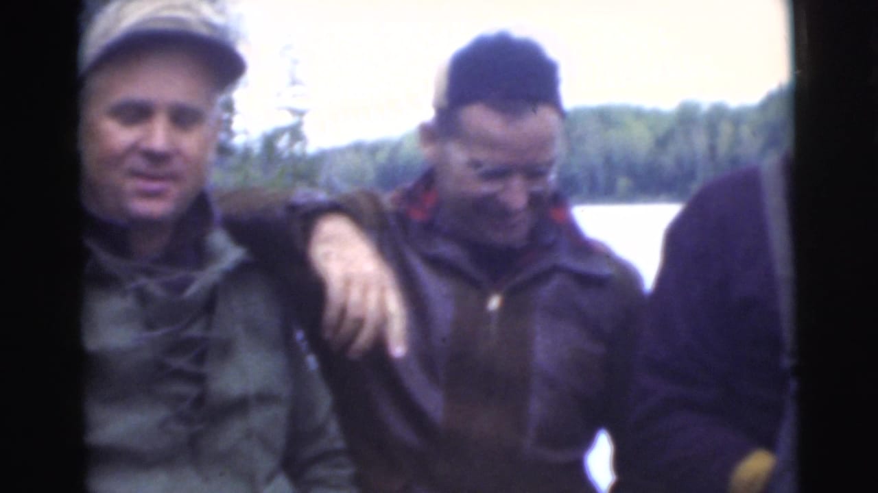 Two men in caps stand close together near a lake and trees.
