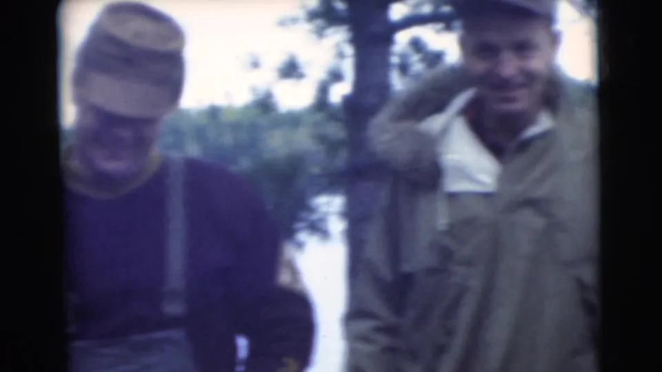 Two adult men in 1950s outdoor clothing stand near lake water, with one wearing suspenders and a cap.