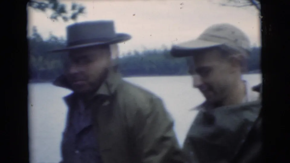 Two adult men wearing hats and outdoor jackets stand beside lake water in a 1956 Miami-labeled 8mm frame.