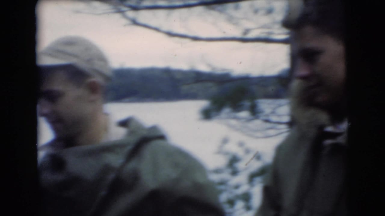 Two men in caps and jackets stand near water and bare tree branches.