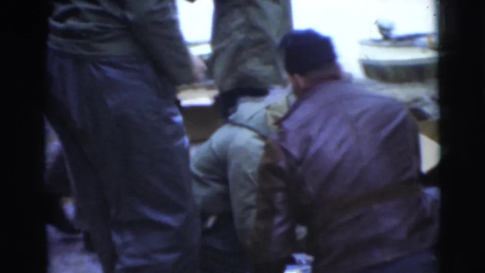 1956 Miami frame showing seated and standing men in jackets around a lakeside outdoor camp meal area.
