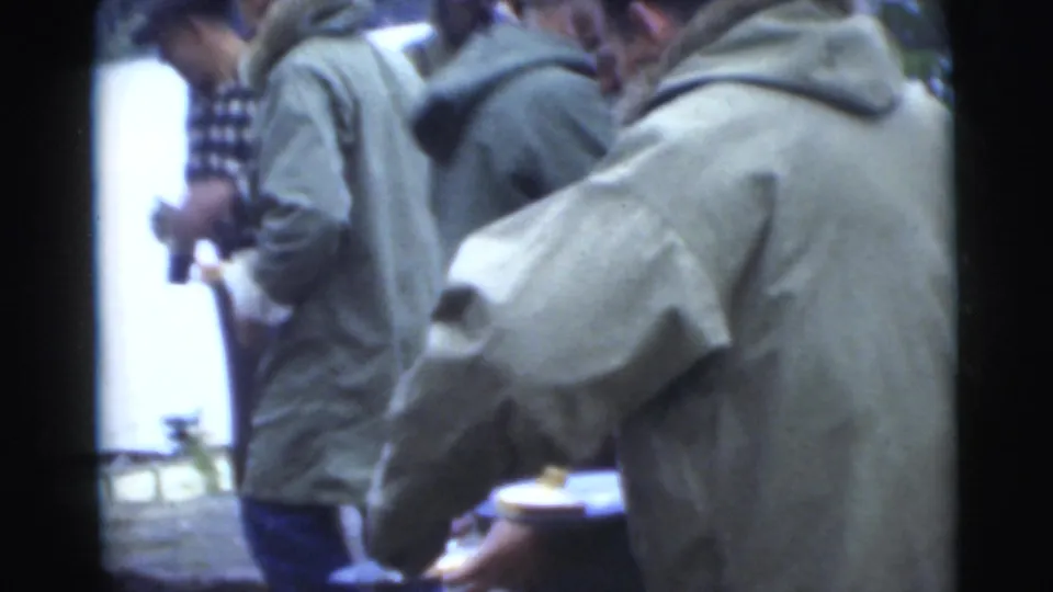 A man with a metal cup stands among other jacketed men near the lakeside camp area in 1956 Miami footage.