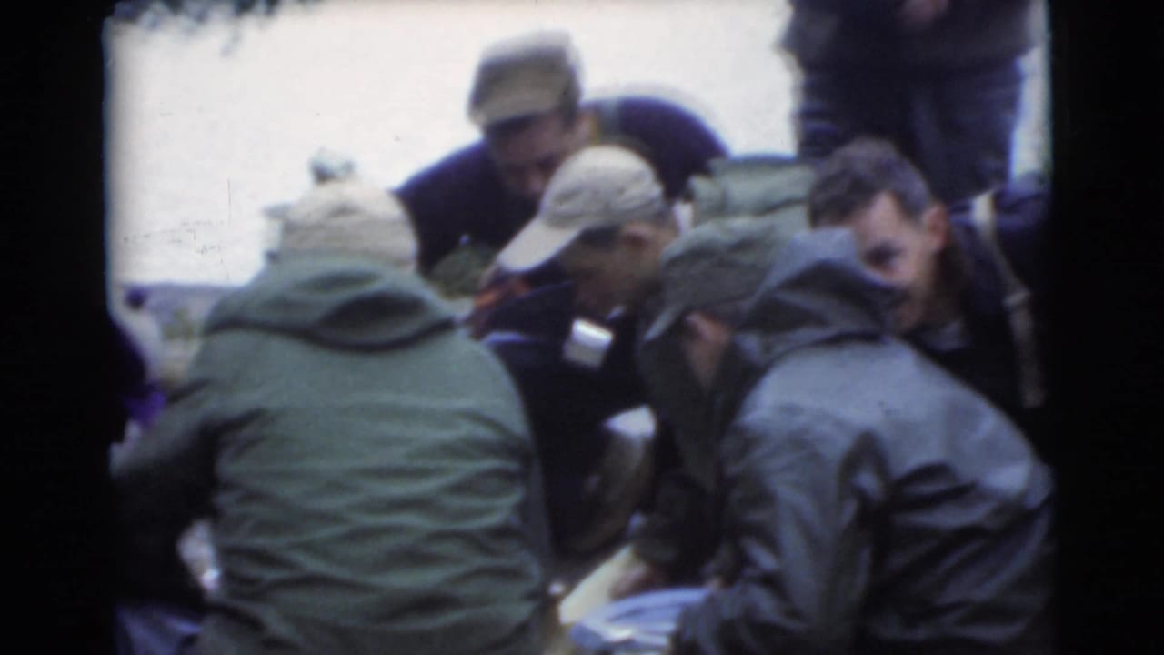 Several men in coats and caps crouch or lean together near water.