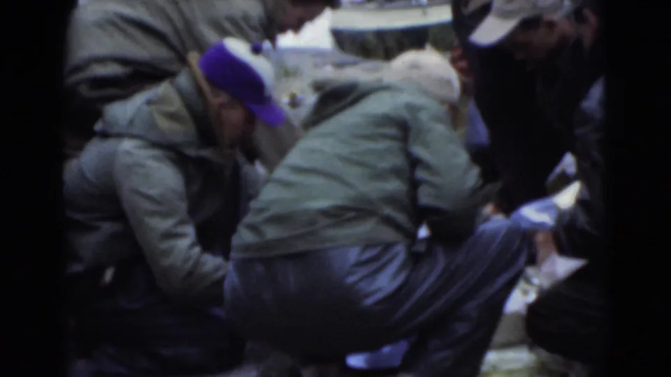 Candid 1956 Miami frame of bundled men crouching near water, with soft focus and visible 8mm film texture.