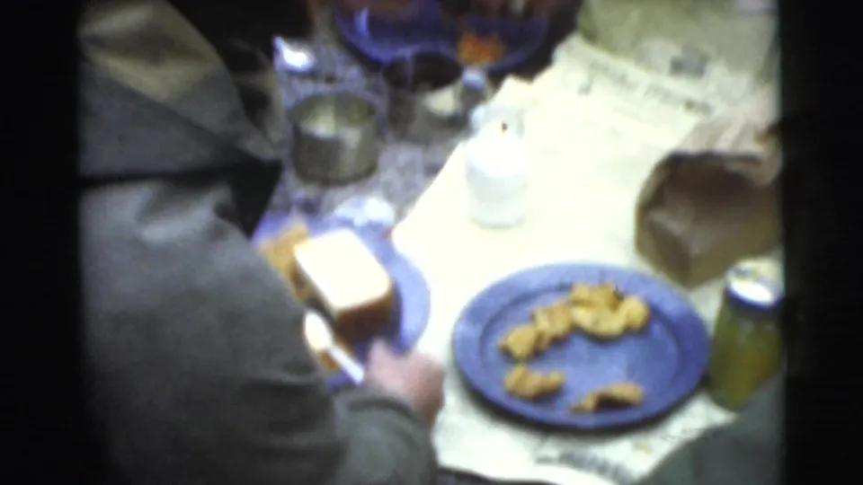 The camera looks down on a small spread of food, dishes, and containers as men gather around in the supplied 1956 Miami footage.