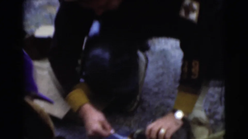 Men in coats crouch closely around food or supplies on rough ground in this dark, handheld 1956 Miami home-movie frame.