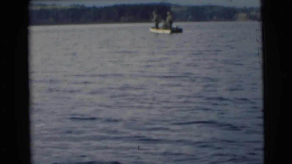 1956 Miami-area 8mm frame showing two uniformed men on a small motorboat against calm water and a dark shoreline.
