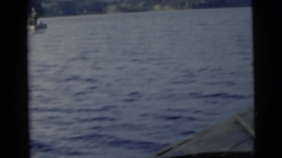 1956 Miami-area 8mm frame with a distant small boat, wide water surface, shoreline, and part of the filming boat visible at lower right.