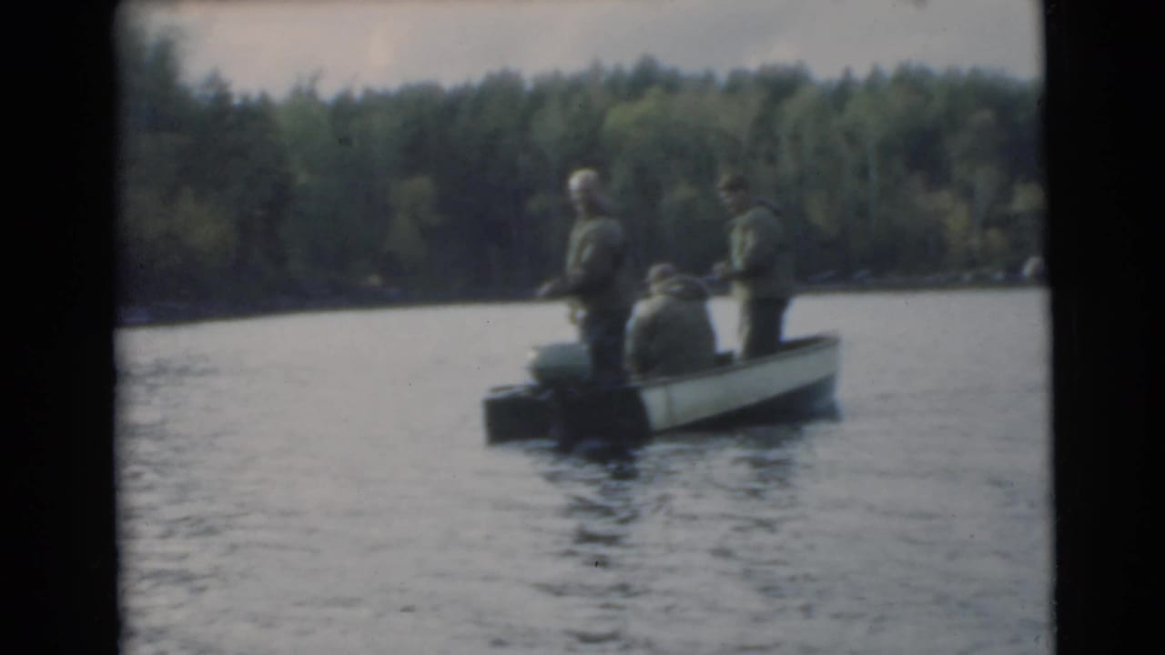 Three men in a small motorboat on a lake near a wooded shoreline.