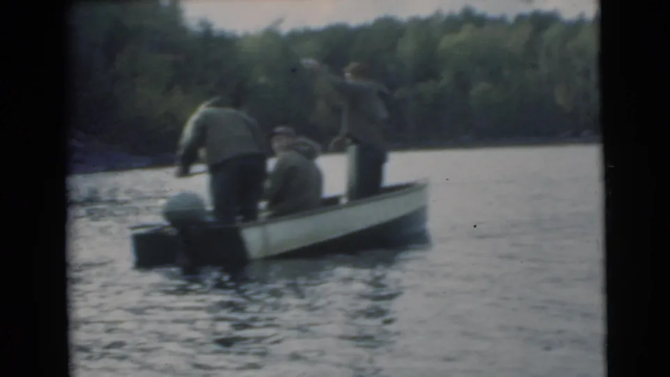 Two men stand and one sits in a small fishing boat on a wooded lake in archival 8mm footage.