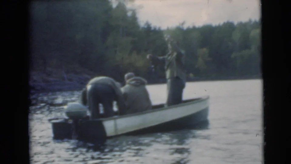 One man raises a fishing rod while two others sit or bend in a small motorboat on a lake near trees.