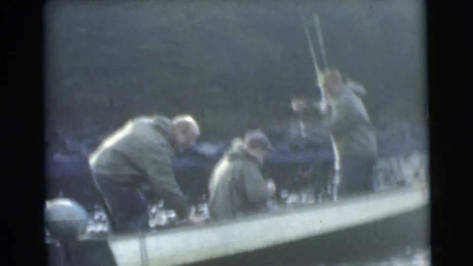 Soft 8mm frame from 1956 Miami showing adult men leaning over a small motorboat during a fishing moment.