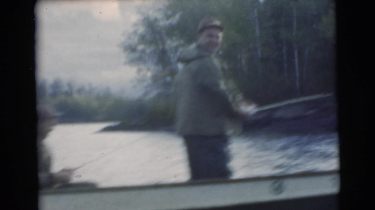 Man standing in a small boat on a lake near a wooded shoreline.