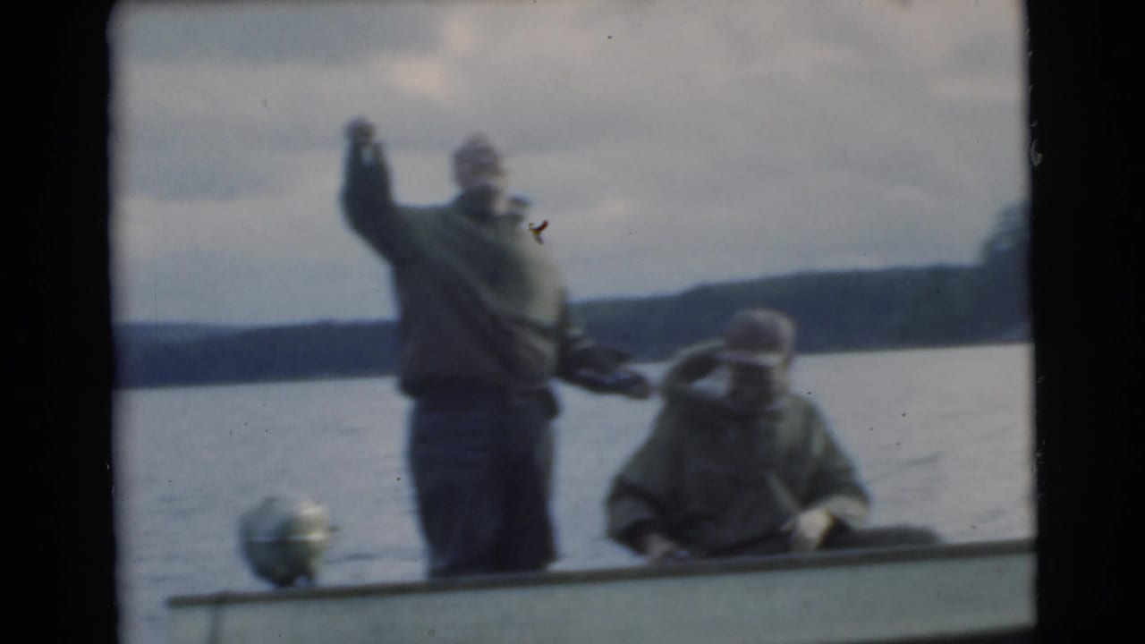 Two men in a small boat, one standing with an arm raised and one seated.