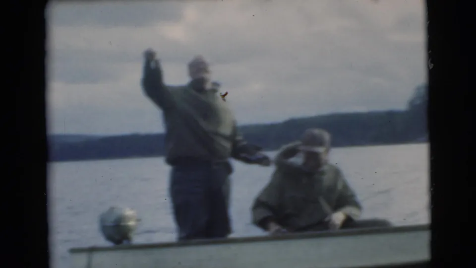 1956 Miami home-movie frame showing two men in a small boat on a lake, one standing with his arm raised and one seated.