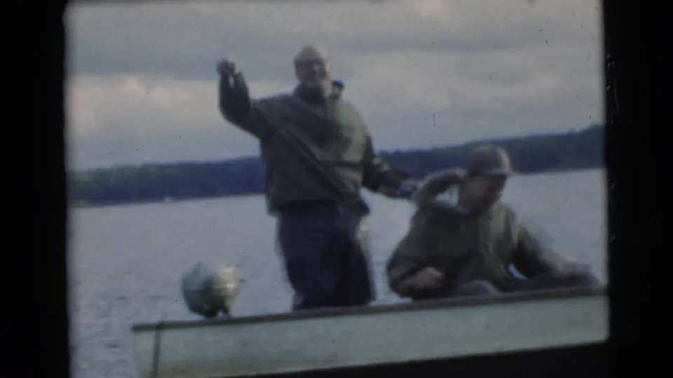 Mid-century 8mm frame from Miami showing two men fishing or handling gear in a small boat on calm lake water.