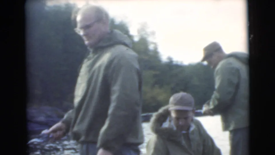 1956 Miami-area home-movie frame with men in hooded jackets and caps in a small boat near a wooded lakeshore.