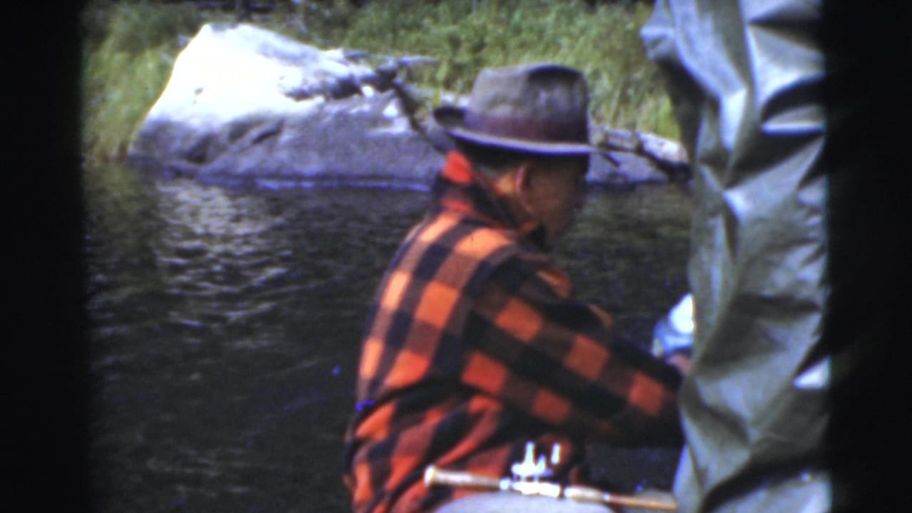 Man in a plaid jacket and brimmed hat sits in a boat beside rippling water.