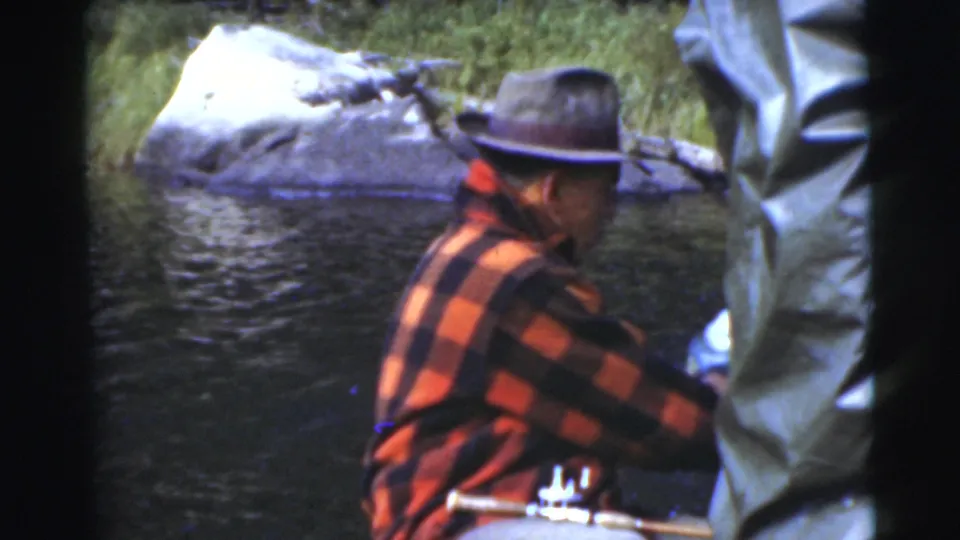 In supported 1956 Miami home-movie footage, a man in a red and black plaid jacket sits in a small boat near water and shoreline rocks.