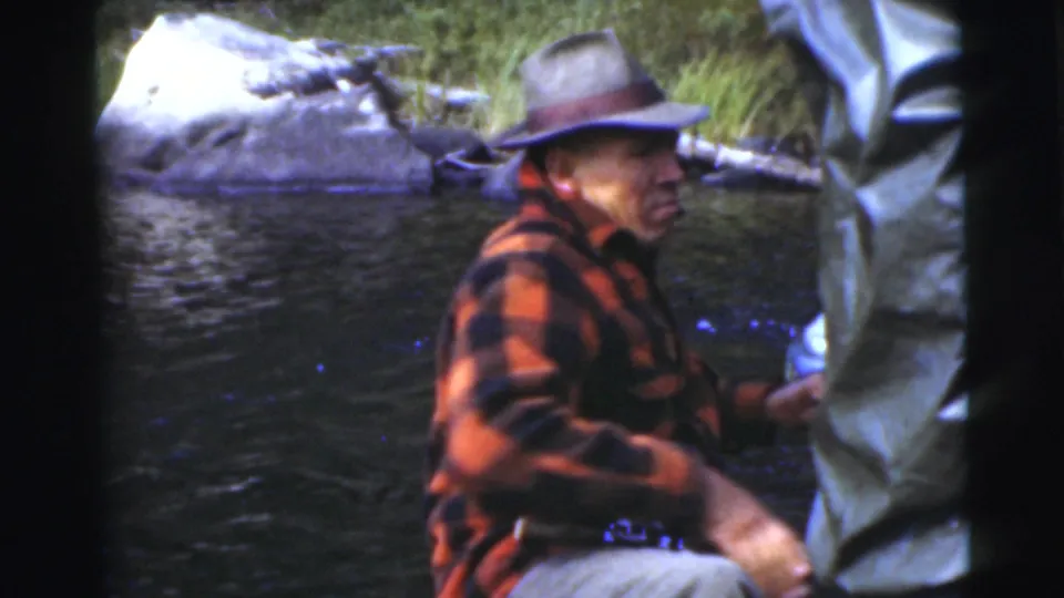 In supported 1956 Miami footage, a seated man in a plaid jacket turns near a standing person in rain gear during a small-boat fishing scene.