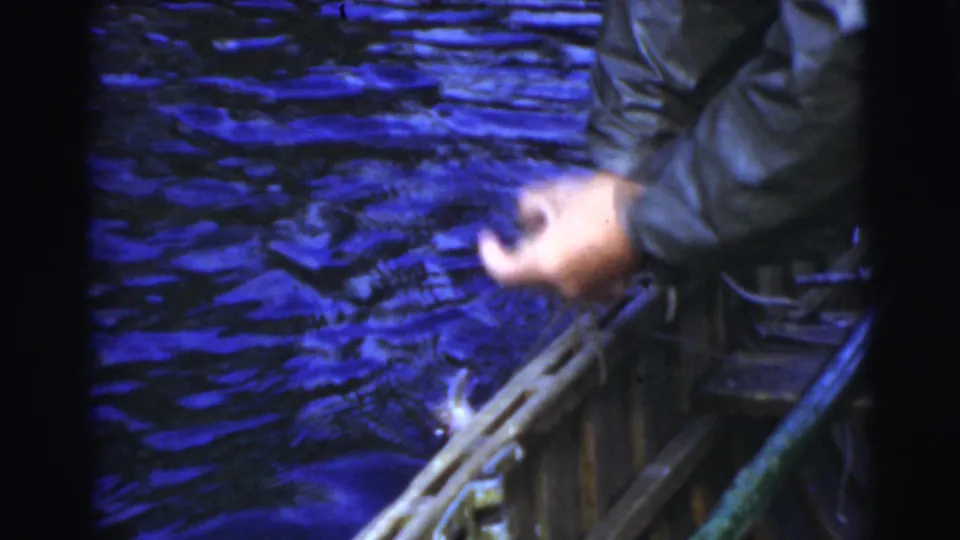 A close 1950s Miami home-movie frame shows a person handling rope or boat gear beside rippling water, with rain gear visible at the edge.