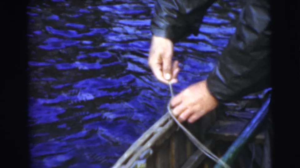 In 1956 Miami home-movie footage, a person's hands pull fishing line from a small boat over dark blue river water.