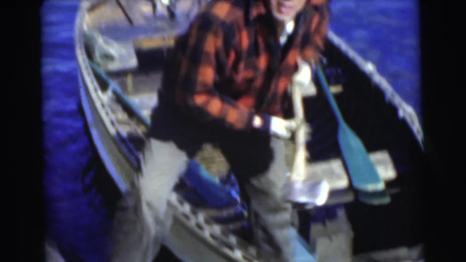Adult man in a red plaid shirt sits in a small rowboat on blue water, with a paddle and handled tool visible.