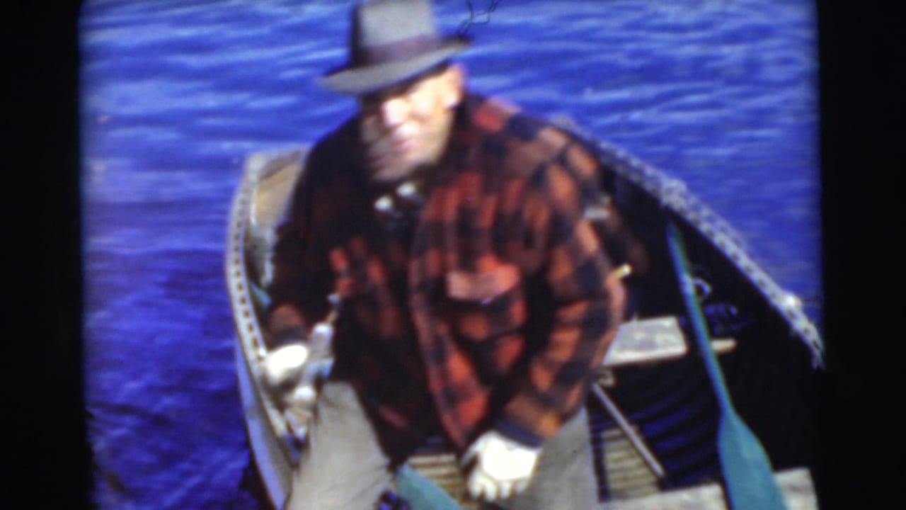 In 1956 Miami home-movie footage, a man in a red plaid flannel shirt and brimmed hat sits in a rowboat on deep blue water.